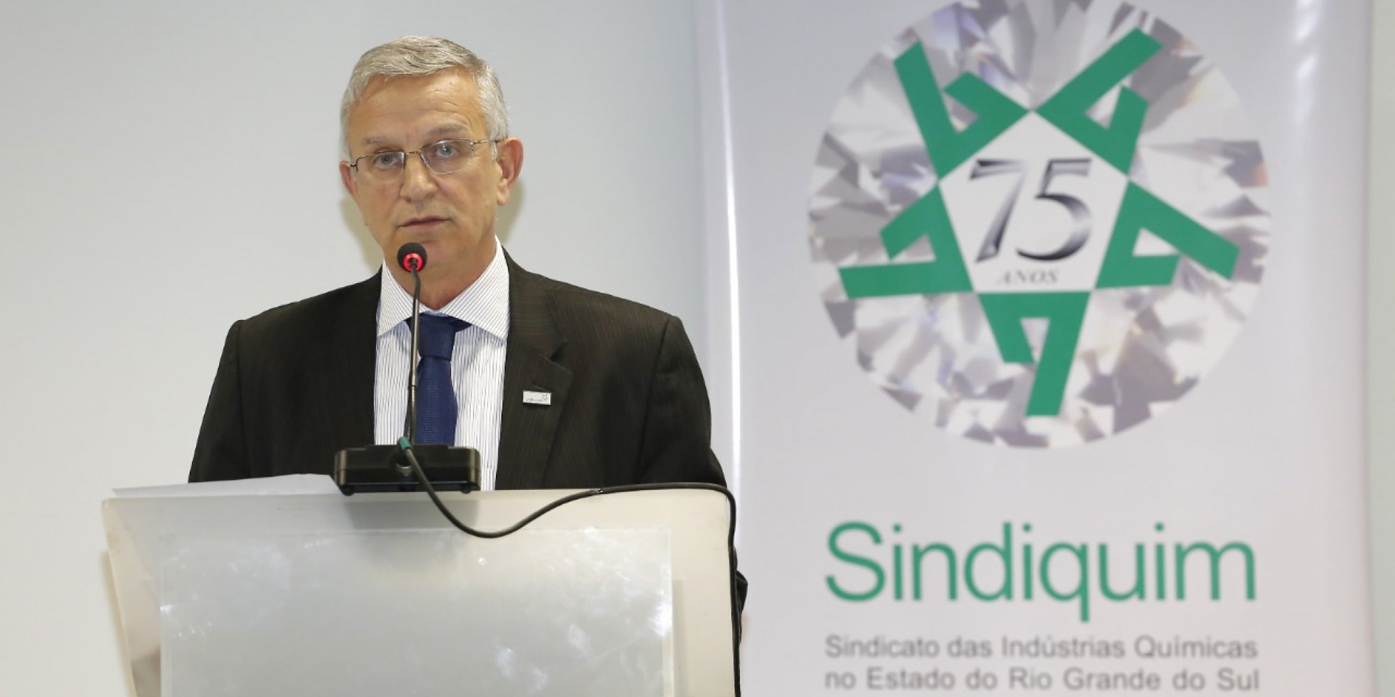 Homem branco, de óculos, em um púlpito com microfone, discursando, banner do Sindiquim Branco escrito em verde